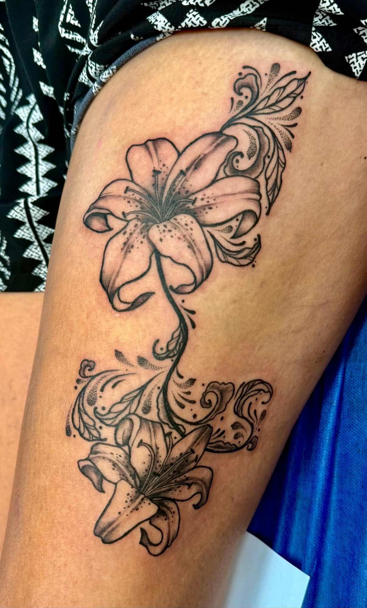 Fine-line lily tattoo with flowing ornamental swirls and dotwork shading.